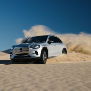 A silver Mercedes GLC drives across a sand dune, throwing up spray