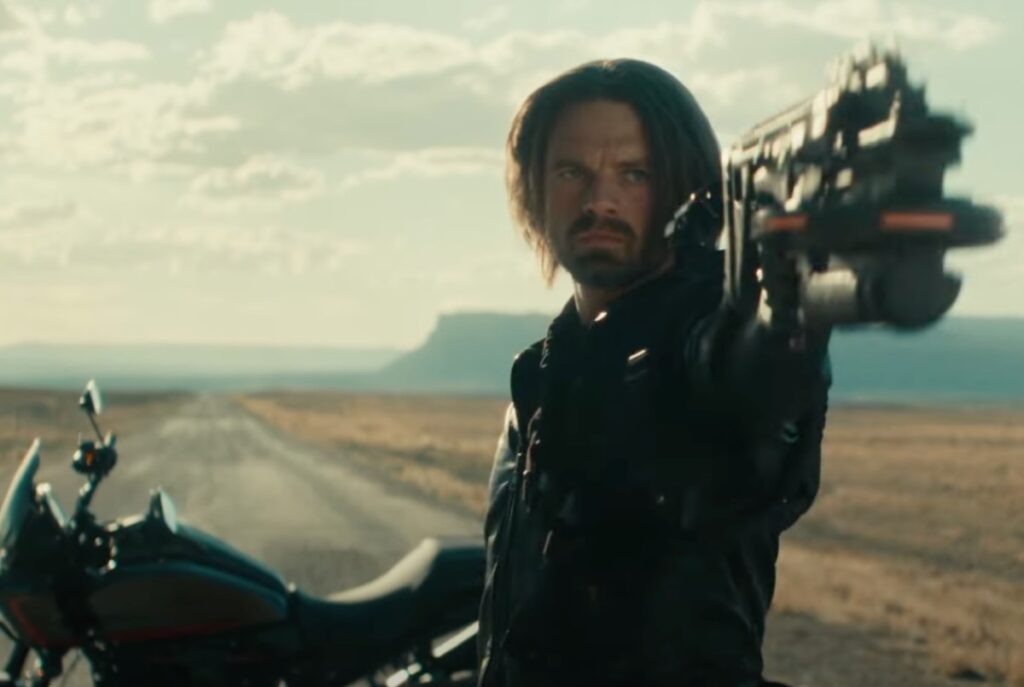 Bearded dark haired man in black outfit pointing a big gun while seated on a motorcycle