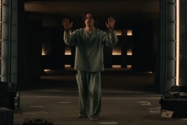 nerdy looking man with brown hair in hospital garb holding up his hands in surrender