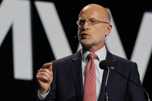 FCC Chairman Brendan Carr delivers a speech on a stage while standing in front of a microphone and gesturing with his hand.