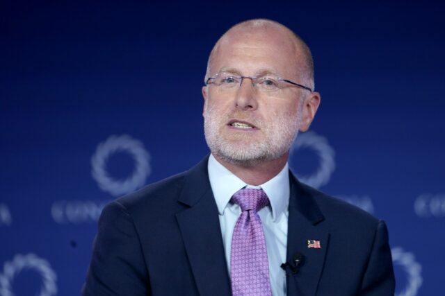 FCC Chairman Brendan Carr speaking on stage at a conference, wearing an American flag pin and a microphone clipped to his suit jacket.