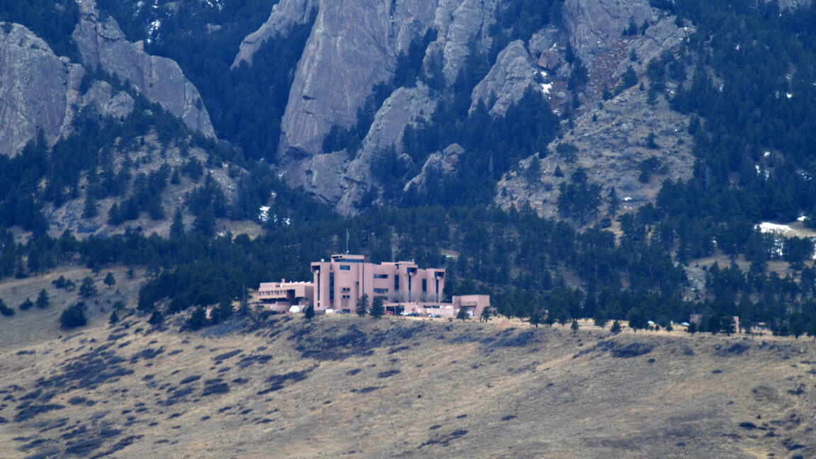 Image of a dry, grassy hill at the foothills of rocky, tree covered mountains. A large cluster of brown buildings sits at the peak of the hill.