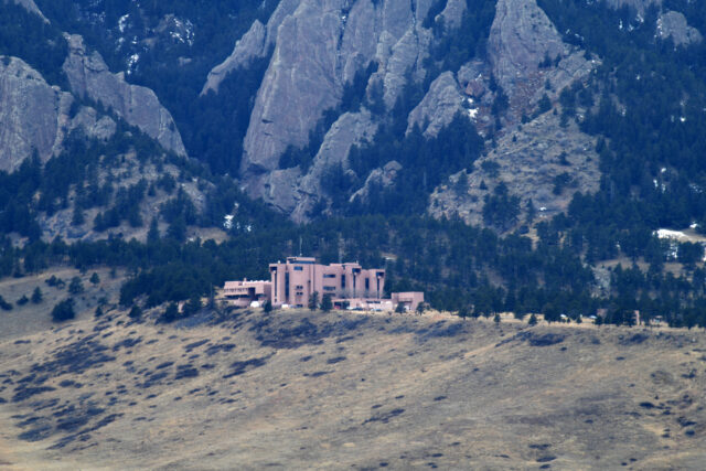 Image of a dry, grassy hill at the foothills of rocky, tree covered mountains. A large cluster of brown buildings sits at the peak of the hill.