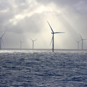 A series of tall wind turbines in the ocean, illuminated by beams of sunlight filtering through baps of heavy clouds.