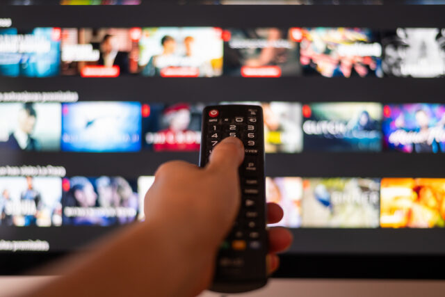 Hand Using A Television Remote Control in front of a TV with a streaming app on display and blurred in the background.