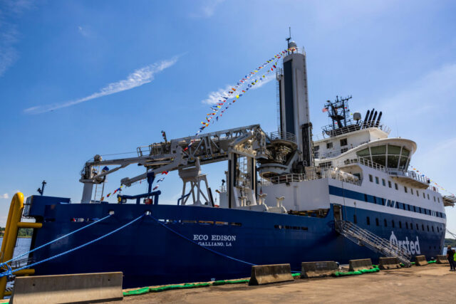 Image of a large blue ship with specialized hardware and cranes on its deck, tied up to a brown dock.