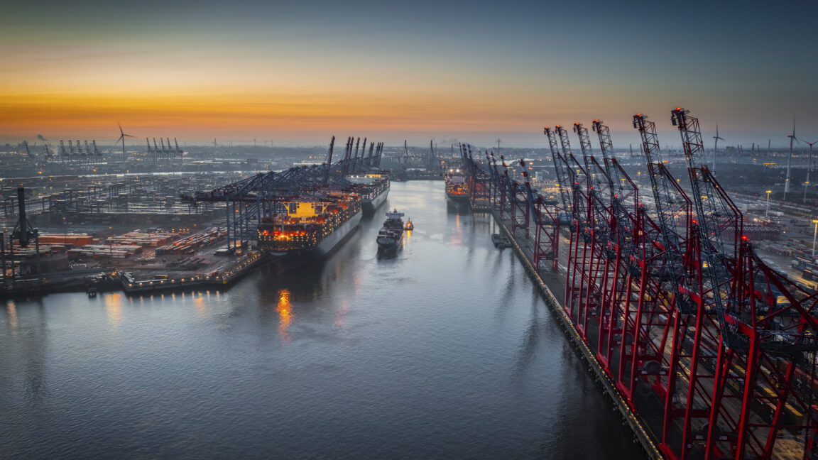 Aerial view of a container terminal with large container cargo ships and gantry cranes. A cargo ship is navigating through the central waterway. On both sides of the waterway are massive container terminals lined with red and black gantry cranes used for loading and unloading ships. on the left docked ships with brightly lit containers. Moody sky at dawn.