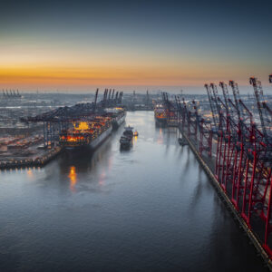 Aerial view of a container terminal with large container cargo ships and gantry cranes. A cargo ship is navigating through the central waterway. On both sides of the waterway are massive container terminals lined with red and black gantry cranes used for loading and unloading ships. on the left docked ships with brightly lit containers. Moody sky at dawn.