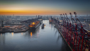 Aerial view of a container terminal with large container cargo ships and gantry cranes. A cargo ship is navigating through the central waterway. On both sides of the waterway are massive container terminals lined with red and black gantry cranes used for loading and unloading ships. on the left docked ships with brightly lit containers. Moody sky at dawn.