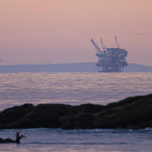 A surfer is paddling in the ocean water as the Hondo offshore oil platform looms in the distance at Refugio State Beach Santa Barbara, California.