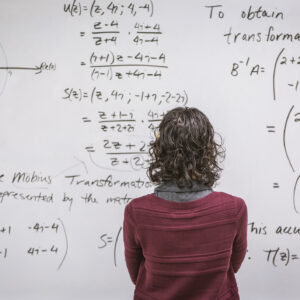 A woman standing in front of a white board that is packed with equations.