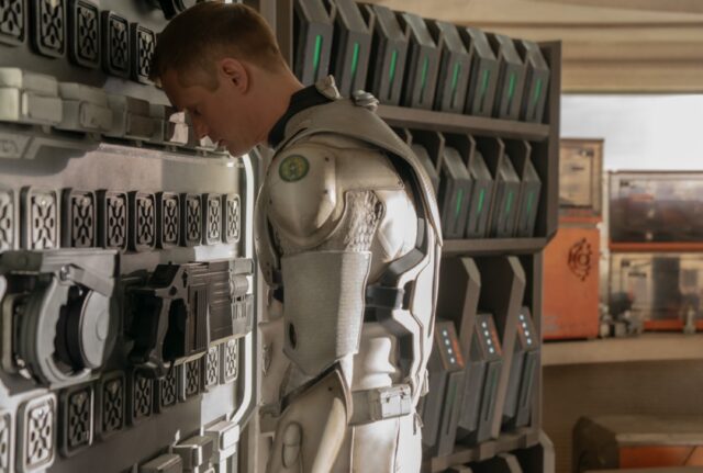 murderbot with fave revealed, standing in a corner with his head bent and leaning against the wall, back to other other people