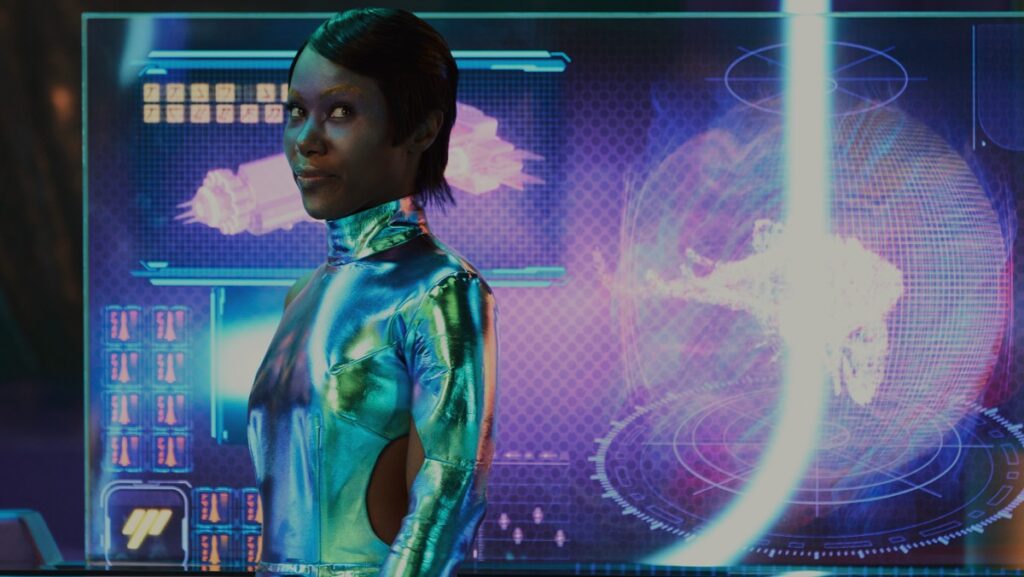 black woman with short hair looking at camera while sanding sideways, in a green-blue body suit.