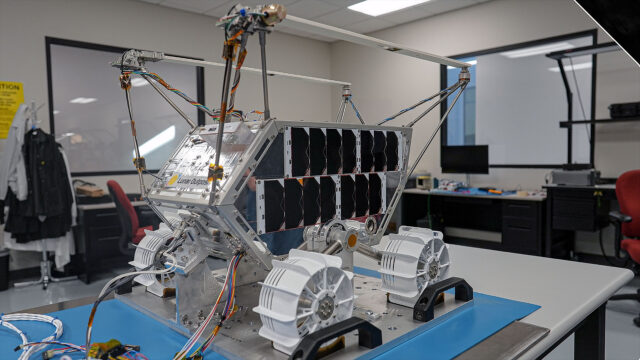 a small, four-wheel lunar rover with solar panels along one side is seen in a lab being prepared for its launch