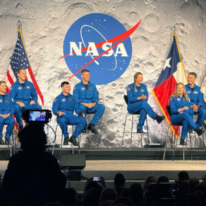 six women and four men, all dressed in blue flight suis, sit on a stage backdropped by images of Earth, the moon and Mars.