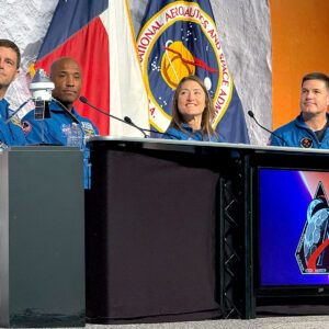 Three men and a woman, all in blue flight suits, sit at a desk during a press conference