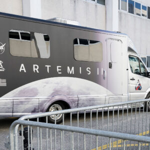 A motor coach decorated with an image of the moon is parked outside of a building.