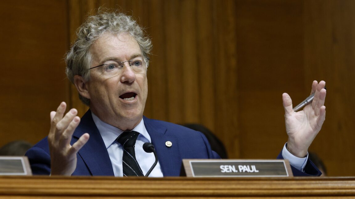 Senator Rand Paul sitting and holding his hands out at his sides while speaking during a committee hearing.