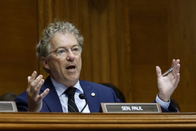 Senator Rand Paul sitting and holding his hands out at his sides while speaking during a committee hearing.