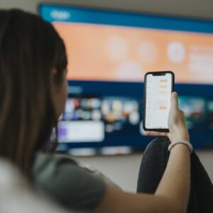 A girl uses a smartphone while watching a TV mounted on a living room wall.