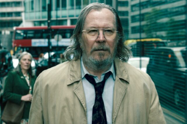 scruffy bearded older man in a beige trenchcoat walking down busy London street