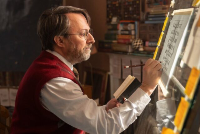 middle aged man in dress shirt and short sleeved sweater meticulously working on a puzzle on an easel